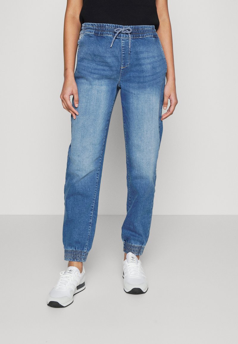 Světle modré joggingové kalhoty z denimu, s páskem na šňůrku, elastickými manžetami a volným střihem. V kombinaci s bílými sportovními botami.