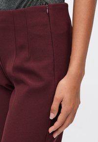 Close-up da mão de uma pessoa repousando perto do fecho das calças burgundy de cintura alta e uma blusa cinza.