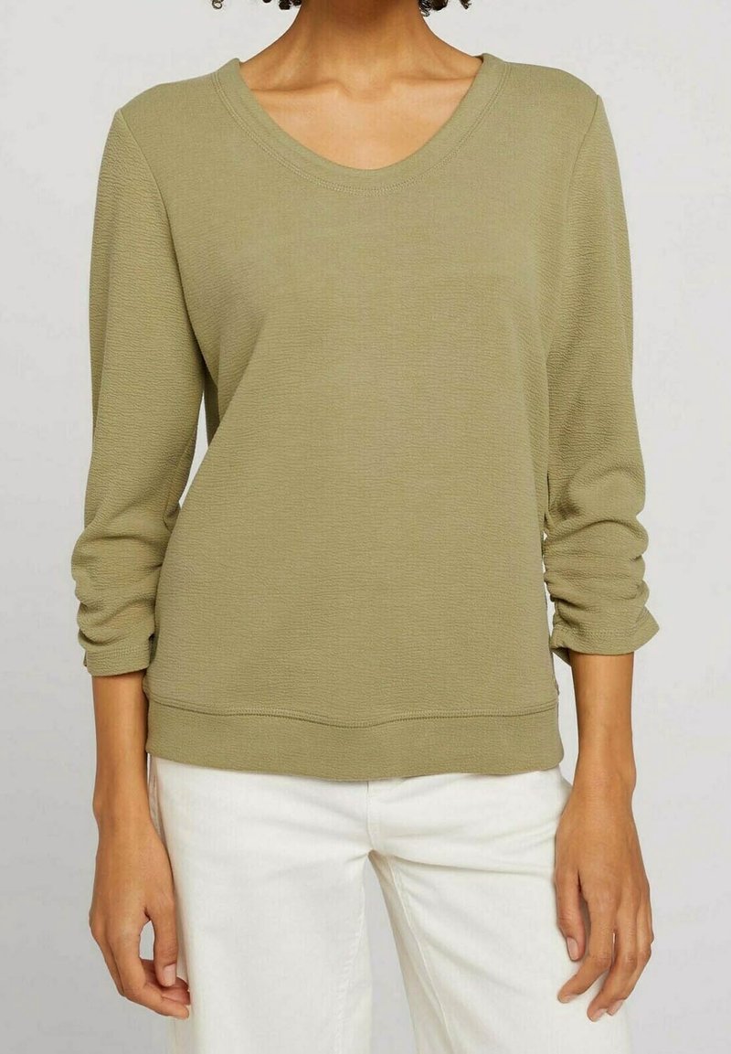 Personne portant un haut texturé vert olive clair à manches longues avec un col rond et un pantalon blanc sur un fond neutre.