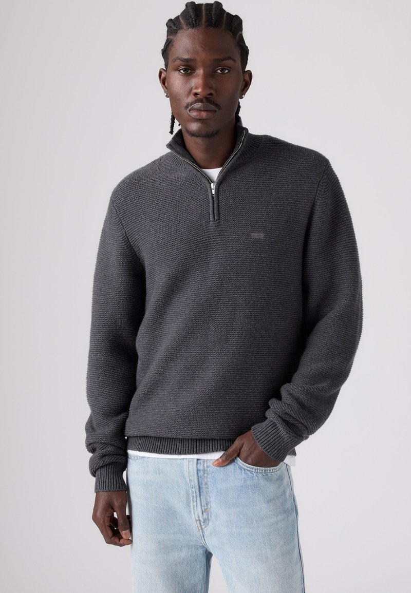 Homme aux cheveux tressés portant un pull gris foncé à fermeture éclair semi-ouverte et un jean bleu clair, debout une main dans la poche devant un fond uni.