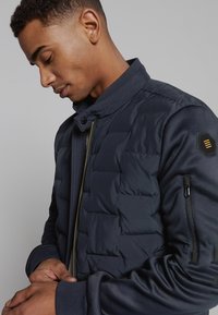 Marineblauwe gewatteerde jas met hoge kraag, geribbelde manchetten en een ritszak op de mouw. Beschikt over een hexagonaal logo-opgenaaide patch op de schouder.