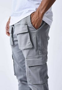 Pantaloni cargo grigi con multiple tasche, una vestibilità affusolata e una texture liscia. Indossati con una t-shirt bianca, che mette in mostra le tasche su entrambi i lati.