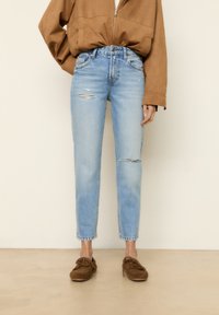 Persona con jeans desgastados de color azul claro, mocasines de ante marrón y una chaqueta marrón con cremallera, con las manos en los bolsillos, de pie sobre un suelo beige.