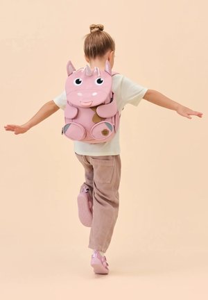Child balancing on one foot with arms extended, wearing a pink unicorn backpack, light shirt, pink pants, and matching shoes against a beige background.