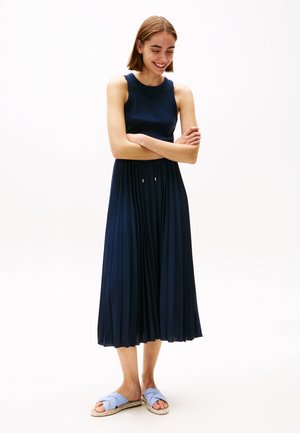Navy pleated dress with a fitted sleeveless top and flowing skirt. Paired with light blue cross-strap sandals on a light background.