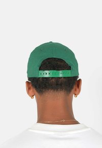 Grüne Baseballmütze mit strukturiertem Oberteil, aus Stoffmaterial und verstellbarem Kunststoffband hinten. Rückwärts getragen, zeigt lockiges Haar.