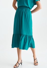 Jupe midi teal en tissu lisse avec taille élastique, dotée d'un ourlet froncé pour plus de détails. Associée à des talons noirs à lanières.