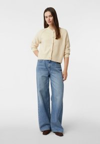 Krótki beżowy kardigan z okrągłym dekoltem i guzikami, zestawiony z szerokimi, jasnoniebieskimi jeansami. Gładka faktura tkaniny, casualowy krój.