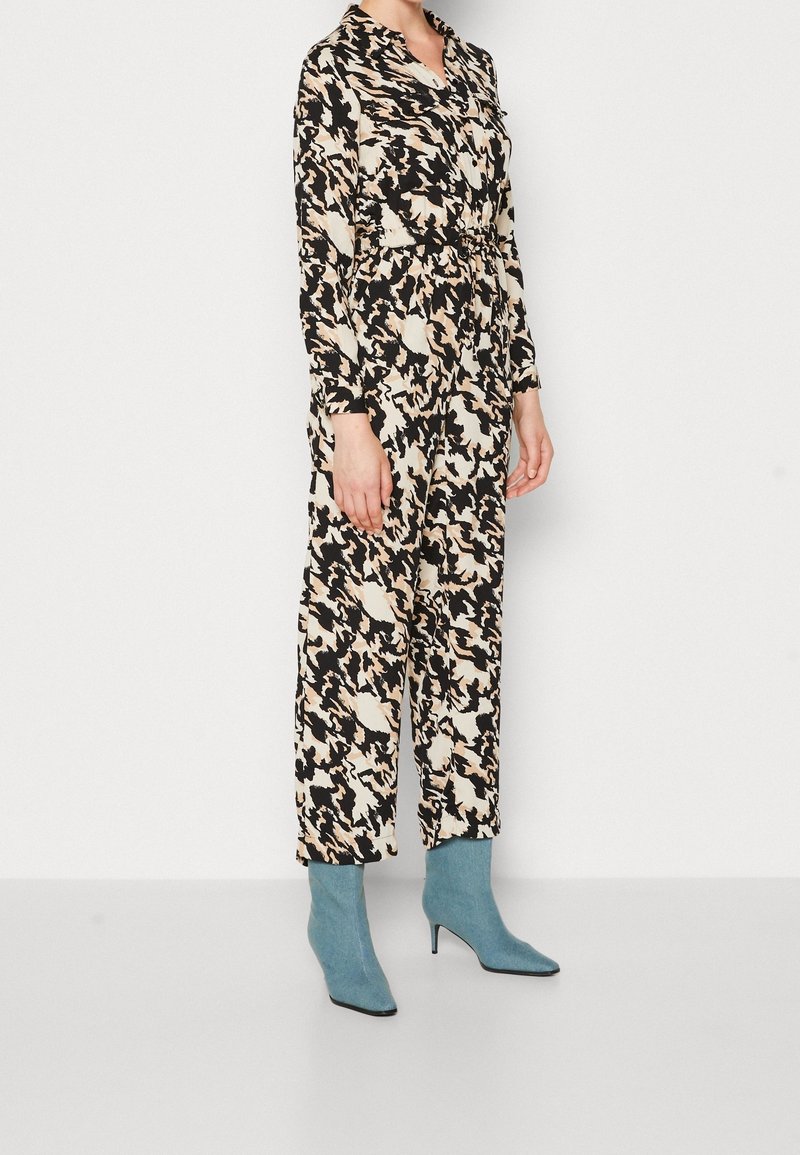 Femme portant une combinaison à manches longues noire et blanche à motifs, associée à des bottines à talons bleu clair, debout sur un fond blanc uni.