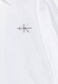 Camicia bianca con bottoni realizzata in cotone, con un logo lavanda "Calvin Klein Jeans" e una stilizzazione di "cK" sull'area sinistra del petto.