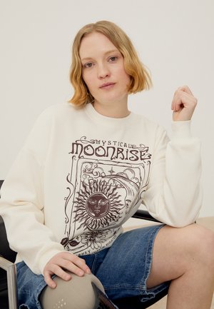 Žena sedí, má na sebe bielu mikinu s nebeským dizajnom slnka a mesiaca a modré rifľové krátke nohavice, pózuje s rukou zdvihnutou blízko tváre.