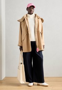 Beige hettejakke med snøring, kremfarget ribbestrikket genser, marineblå vid bukser, rosa caps og hvite joggesko. Holder en beige totebag.