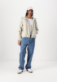 Light beige hooded jacket with elastic cuffs, white t-shirt, loose blue jeans, and white sneakers with brown accents. Casual outfit.