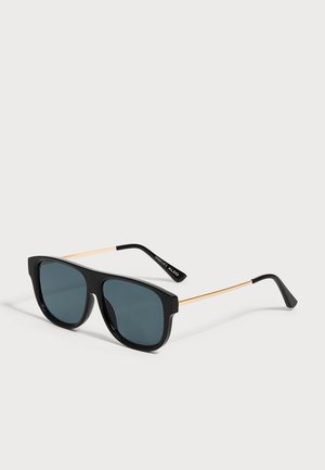 Lunettes de soleil carrées noires avec verres foncés et fines branches métalliques dorées, reposant sur une surface grise neutre.