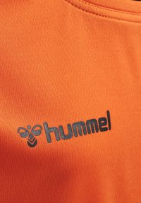 Oranger Stoff mit strukturierter Oberfläche zeigt ein schwarzes "hummel"-Logo und ein Bienenmotiv, das den sportlichen Stil der Bekleidung unterstreicht.