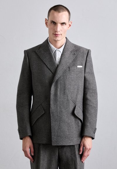 Veste double boutonnage en mélange de laine grise avec de grandes poches angulaires, des revers en pointe et des poignets à boutons. Détail de l'étiquette blanche sur la poche poitrine.