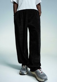 Sort corduroy bukser med en løs pasform, der har lodret ribbet tekstur, sidelommer, båret med grå sportsko. Hvid t-shirt synlig.