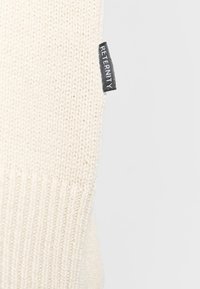 Knit sweater in off-white with ribbed texture. Features a small black label with "RETERNITY" attached at the side.
