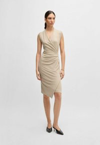Beige sleeveless wrap dress with a deep V-neck, draped texture, and asymmetrical hemline. Paired with black heeled mules.
