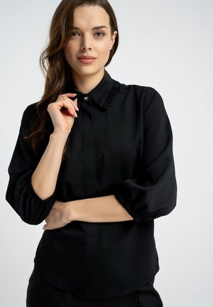 Kobieta z falującymi brązowymi włosami, ubrana w czarną bluzkę z długim rękawem i zapiętym kołnierzykiem, stojąca na tle jednolitego jasnego tła.