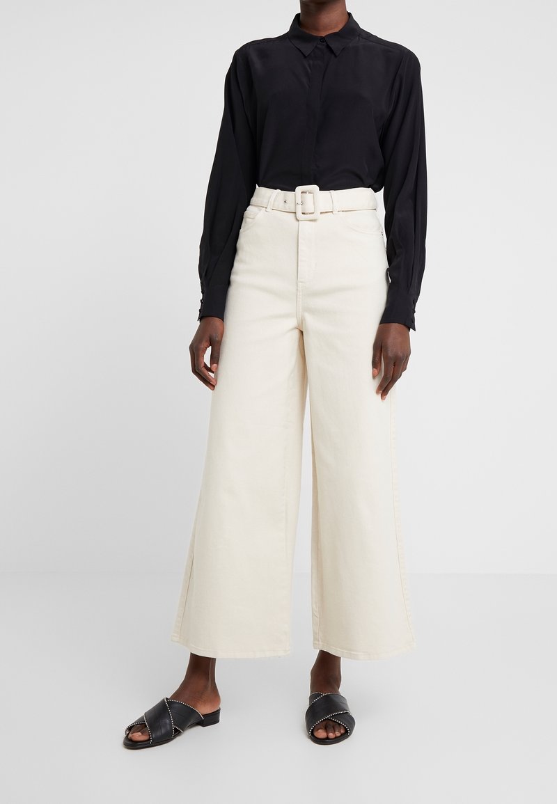 Czarna koszula z długim rękawem na guziki, zestawiona z kremowymi szerokimi spodniami z paskiem. Czarne klapki z krzyżującymi się paskami i ozdobione ćwiekami.