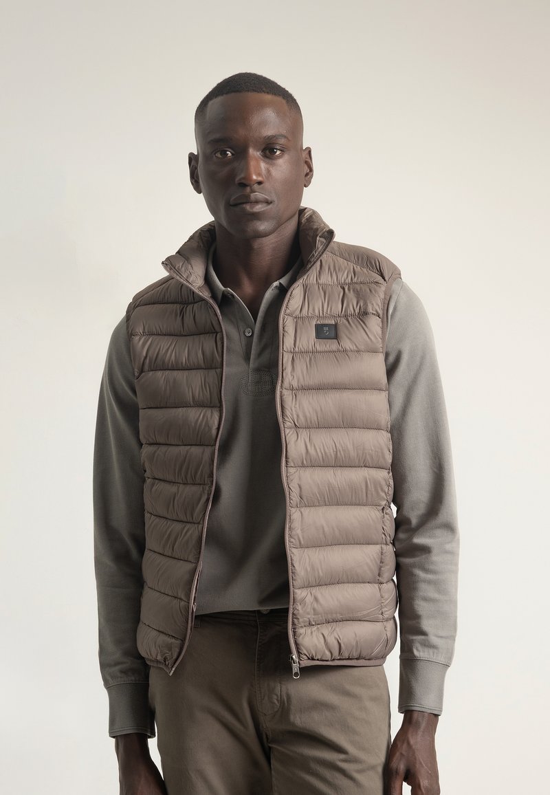 Chaleco acolchado ligero en color beige con un cuello alto, costuras verticales y cierre de cremallera. Llevado sobre una camiseta de manga larga gris.