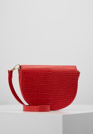 Rød håndtaske formet som en halvmåne med krokodillestruktur og aftagelig rem, vist på en hvid piedestal mod en ensfarvet baggrund.