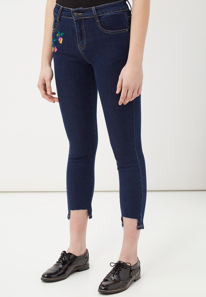 Jeans de denim azul oscuro con un corte ajustado, que presentan bordados florales en la cadera izquierda y dobladillos deshilachados en los tobillos. Combinados con zapatos de charol negros.