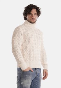 Maglione a collo alto in maglia cable-knit crema con polsini e orlo a costine, caratterizzato da un motivo texturizzato. Abbinato a jeans blu strappati.