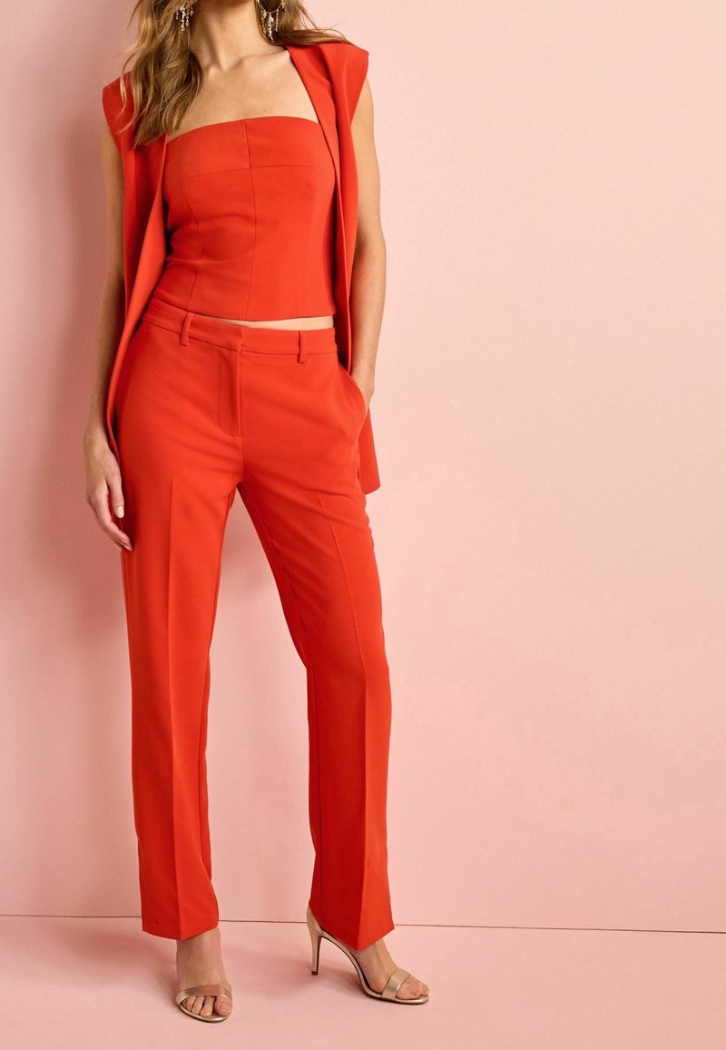 Red tailored pantsuit featuring a strapless top, slim-fit trousers, and a sleeveless blazer. Smooth fabric with a solid color, minimal accents.