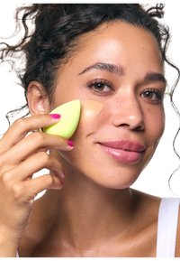 Kobieta z kręconymi włosami nakłada podkład na policzek za pomocą limonkowozielonej gąbki do makijażu, lekko się uśmiechając, w białej bluzce.