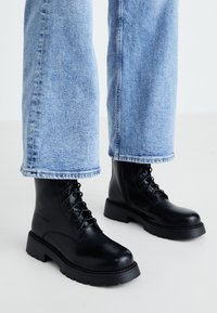 Botine din piele neagră, cu vârful rotund, cu șireturi în față și o talpă groasă, texturată. Pur state cu pantaloni din denim de culoare albastru deschis.