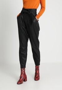 Černé kalhoty z umělé kůže s vysokým pasem, širokým opaskem a bočními kapsami, doplněné lesklými červenými kotníkovými botami se vzorem hadí kůže.