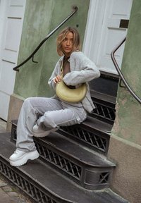 Borsa gialla rotonda, cardigan grigio, top marrone, pantaloni a righe e scarpe sportive bianche spesse. Seduta su gradini neri a motivo, sfondo verde.