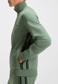 Žalios spalvos užsegamas džemperis iš minkštos medžiagos, su aukštu kaklu, šoninėmis kišenėmis ir juodomis vertikaliomis juostomis rankovėse.