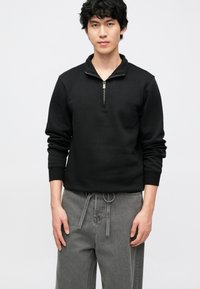 Musta vetoketjullinen collegepaita, jossa on sileä kangas, pystypiha ja resorit hihansuissa, yhdistetty harmaisiin, nauhalla kiristäviin housuihin.