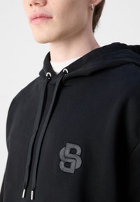 Gros plan d'une personne portant un sweat à capuche noir avec cordons et un logo « SB » entrelacé cousu sur la poitrine.