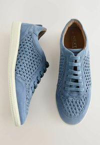 Plave sneakers od nubuka s tkanom teksturom, bijelim gumenim potplatom i šestim rupama za vezivanje. Unutrašnjost ima bež uložak s logotipom branda.
