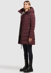 Femeie îmbrăcată într-o paltonă lungă, matlasată, de culoare burgundy, cu glugă, leggings negri și cizme groase negre, stând pe un fundal simplu, de culoare deschisă.