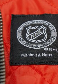 Schwarzes Stoffetikett mit dem NHL-Logo, "Offiziell lizenziertes Produkt" und "Mitchell & Ness" in Weiß gedruckt, vor einem orangefarbenen gesteppten Hintergrund.