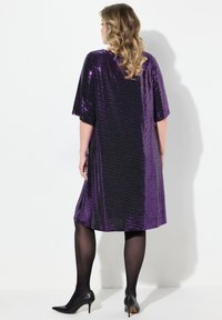 Robe pailletée violette à coupe ample, à manches courtes et longueur genoux. Portée avec des collants noirs et des chaussures à talons.
