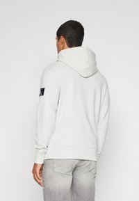 Ljusgrå hoodie av mjukt tyg, med en känguruficka och logotypapplikation på vänster ärm, samt ribbade muddar.