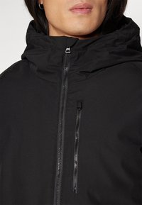 Svart zip-jacka med hög krage, som har en bröstficka med dragkedja och en slät, matt yta.