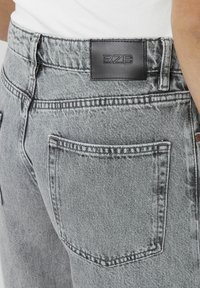 Jeans en denim gris avec une patch en cuir noir portant l'inscription "BZB" à la taille et des poches arrière classiques avec des coutures visibles.