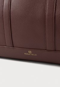 Hnedá kožená kabelka s textúrou, zlatými kovaniami a logom "ANNA FIELD"; má vertikálne panely a štruktúrovaný dizajn.