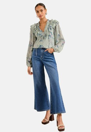 Bluse mit Blumenmuster in hellgrün und Rüschendetails, kombiniert mit weiten blauen Jeans und braunen Plattformsandalen.