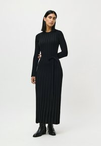 Nainen, joka wears musta ribattua pitkähihaista maksimekkoa, jossa on vyötärösolki, sekä mustia nilkkureita, seisoo yksinkertaista vaaleaa taustaa vasten.