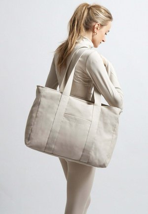 Beige Leinentasche mit dicken Stoffträgern und einer Vorderseite, die eine dezente, in Weiß gedruckte Logo zeigt. Die Tasche hat eine strukturierte Form.