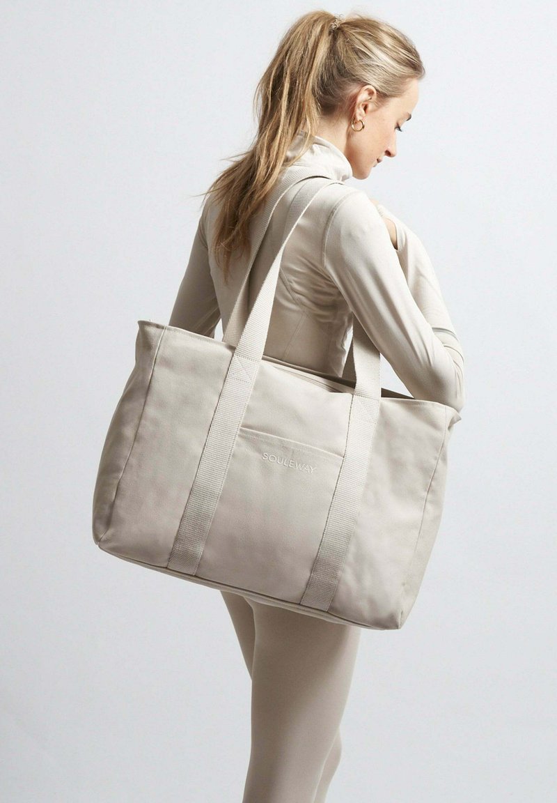 Beige plátěná taška s tlustými popruhy a přední kapsou, na které je vytištěno jemné logo bílou barvou. Taška má strukturovaný tvar.