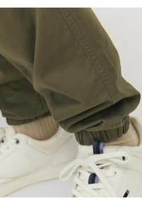 Olijfgroene katoenbroek met elastische boorden, gecombineerd met witte sneakers met blauwe en marine accenten, en geribde beige sokken.